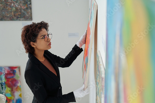 Foto Young gallery owner in white gloves hanging paintings on the wall in gallery