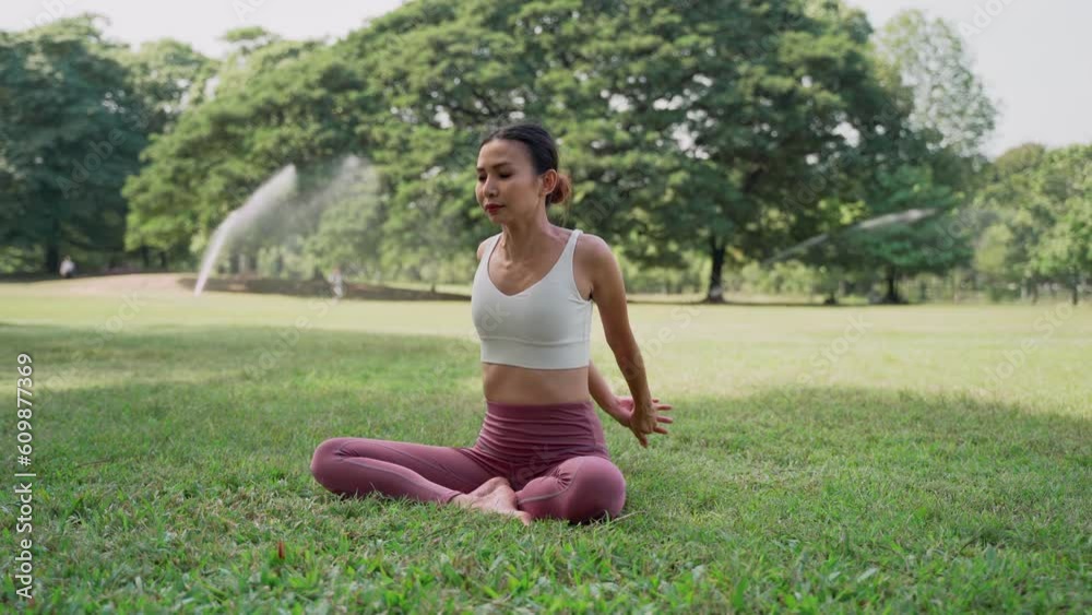 Asian young woman sitting on the grass in the lotus position and raising hands up outside in city park with the big trees background. Rearview of female practicing yoga outdoors on a sunny day. Asian young woman sitting on the grass in the lotus position and raising hands up outside in city park with the big trees background. Rearview of female practicing yoga outdoors on a sunny day.