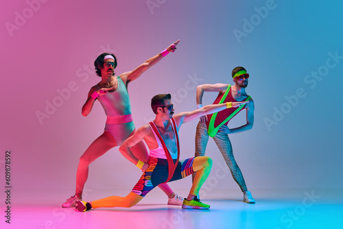 Fototapeta Naklejka Na Ścianę i Meble -  Funny image of three men in stylish, vintage sportswear training aerobics and gymnastics against gradient blue pink studio background. Concept of sportive and active lifestyle, humor, retro style. Ad