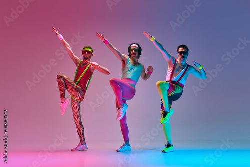 Fototapeta Naklejka Na Ścianę i Meble -  Athletic, funny men in retro colorful sportswear training, doing exercises against gradient blue pink studio background in neon light. Concept of sportive and active lifestyle, humor, retro style. Ad