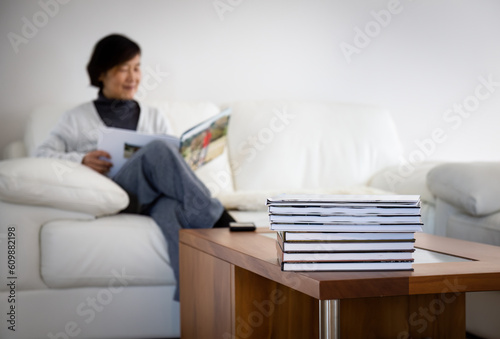 Senior woman smiling and holding family photo album. Selective focus on a pile of photobooks on coffee table.