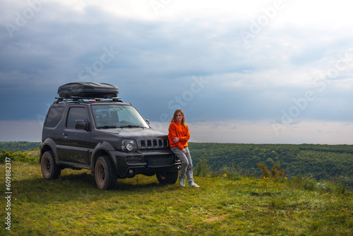Fototapeta Naklejka Na Ścianę i Meble -  A happy girl in a bright orange sweater, is standing next to a small SUV with a roof rack. View point on the top of the mountain, Dniester Subich river, Ukraine. Copy space