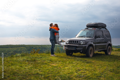 Obraz na płótnie Happy couple kissing and hugging standing on the top of the mountain. Car trip on SUV with roof rack, trip at summer day. Man and woman enjoying time together travelling by vehicle. Copy space w salonie