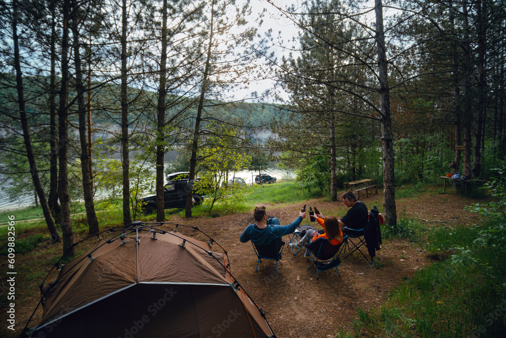 Happy campers. Rearview shot of three friends drink beer, clink bottles. Have fun spending time in nature. Quick folding tent equipment, lightweight chairs. Good mood. SUV off road vehicles