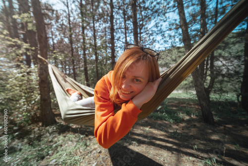 Portrait of a young woman, close up, resting in a hammock in the middle of the forest, summer day. Bright orange sweatshirt. Has a good mood