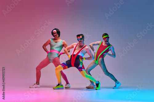 Fototapeta Naklejka Na Ścianę i Meble -  Funny men in vintage, colorful sportswear posing, doing aerobics exercises against gradient blue pink studio background in neon light. Concept of sportive and active lifestyle, humor, retro style. Ad