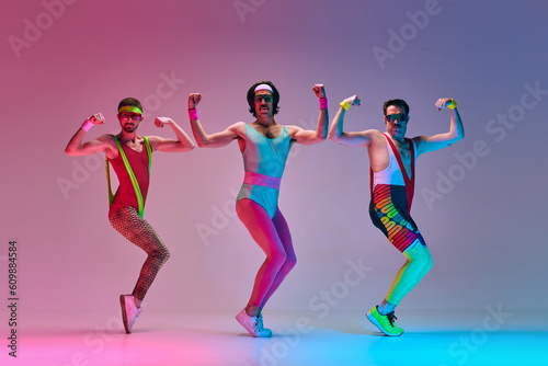 Sport show. Three funny men in colorful sportswear doing aerobics exercises against gradient blue pink studio background in neon light. Concept of sportive and active lifestyle, humor, retro style. Ad photo