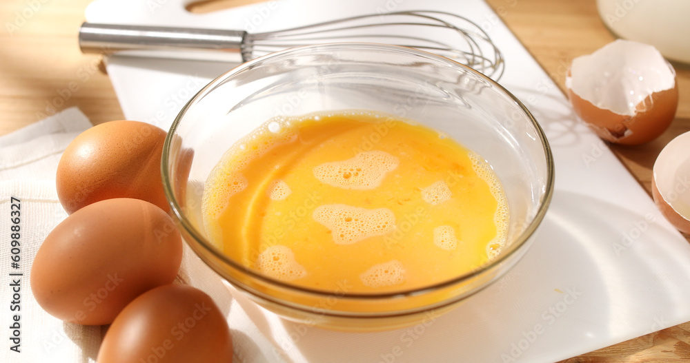 Whisking eggs. Whipped raw eggs in a bowl with whisk. Breakfast preparation. Omelet recipe. Rustic style. Sunny day. Close up. Whisking eggs. Whipped raw eggs in a bowl with whisk. Breakfast preparation. Omelet recipe. Rustic style. Sunny day. Close up.
