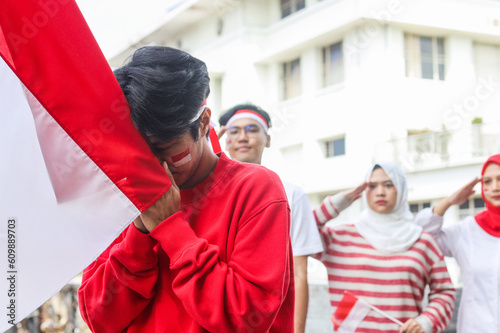 Young Asian man holding and kissing indonesia flag to showing respect while others are saluting to the flag in the background. Indonesian independence day.