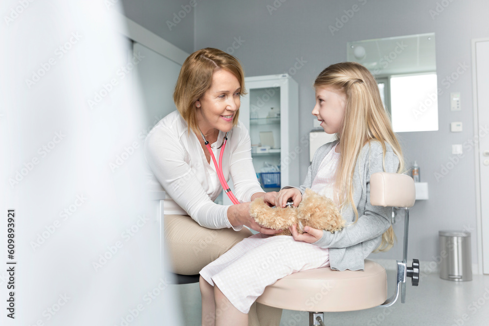 Fototapeta premium Pediatrician using stethoscope on girl patient‚Äôs teddy bear in examination room