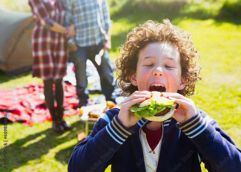 Enthusiastic boy taking large bite of hamburger at sunny campsite Enthusiastic boy taking large bite of hamburger at sunny campsite