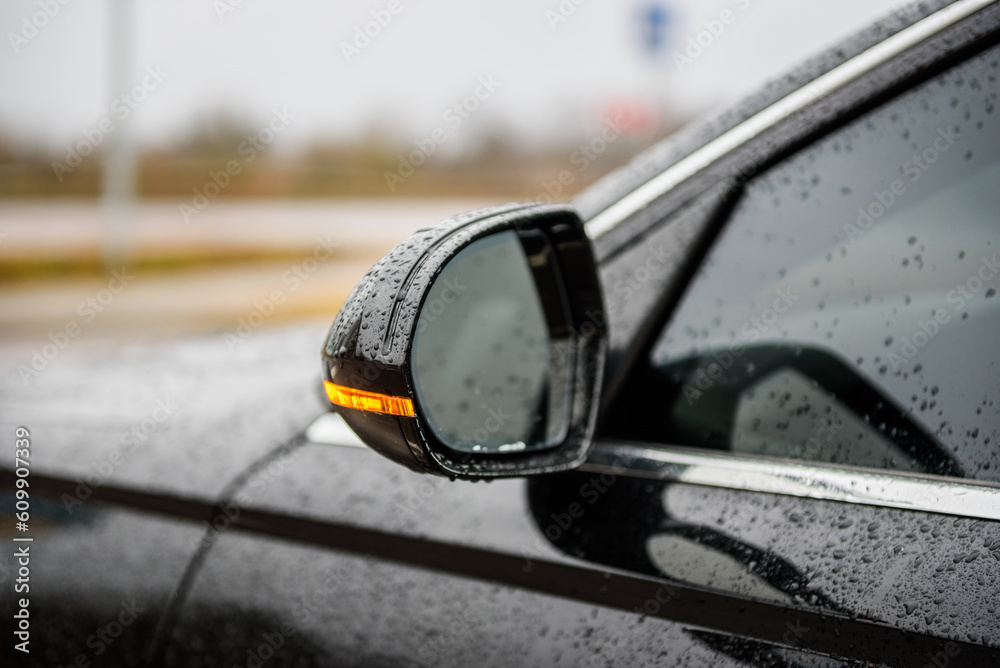 Obraz premium Left rear view mirror in a black car. Side left mirror with turn signal repeater and window of car on street parking and detailing in auto service industry. Road safety while driving. Closeup details.