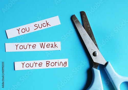 Scissors on blue background with paper pieces written YOU SUCK, You're boring, You're Weak - verbal abuse - negative demeaning words abuser uses to gain or maintain power and control over victims