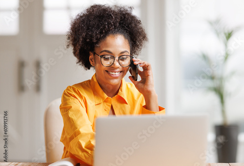 Beautiful young smiling ethnic woman making call via smartphone while working remotely from home while sitting at desk and talking to coworkers . © JenkoAtaman