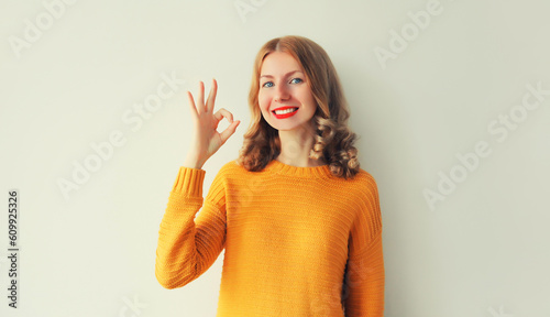 Portrait of happy smiling young woman shows gesture her hand okay sign wearing yellow knitted sweater on white background