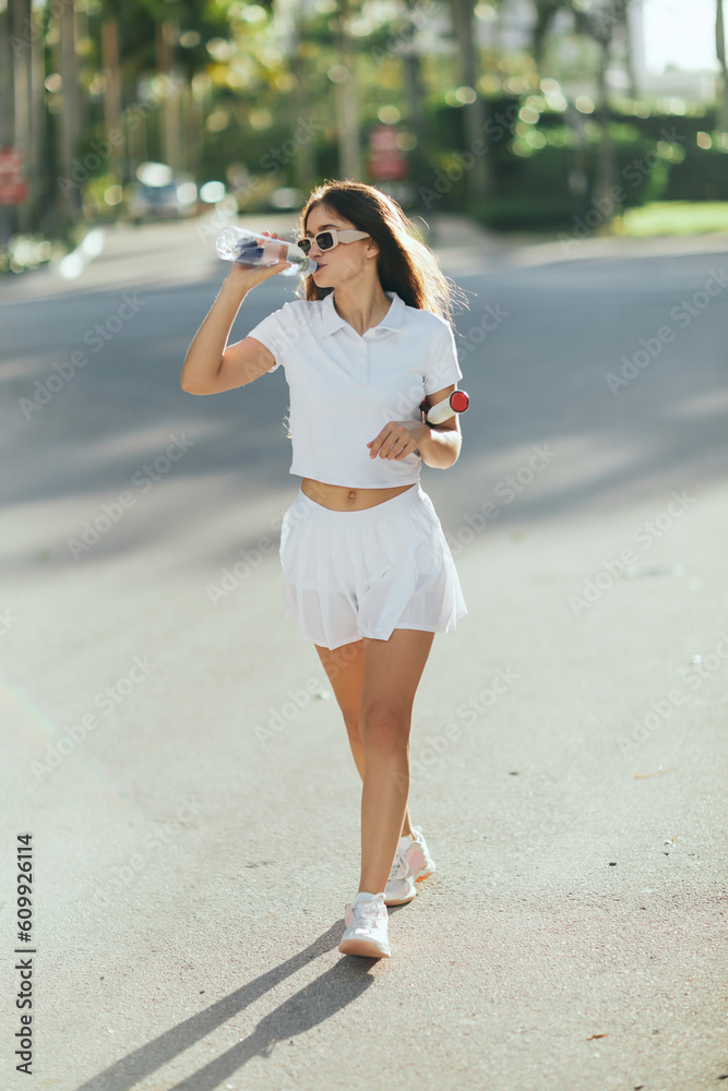 Fototapeta premium female tennis player drinking water, young woman with long brunette hair walking in white sporty outfit and holding racket on urban street in Miami, blurred background, healthy habits