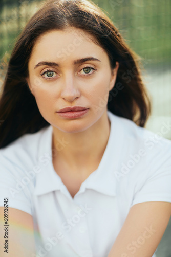 portrait of young woman with brunette hair wearing white polo shirt and looking at camera after training on tennis court, tennis net on blurred background, Miami, Florida, close up