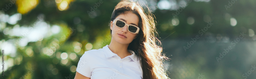 charming and young woman with brunette long hair standing in white polo shirt and trendy sunglasses near blurred and green palm trees in Miami, sunny day, travel, vacation, banner, sun-kissed charming and young woman with brunette long hair standing in white polo shirt and trendy sunglasses near blurred and green palm trees in Miami, sunny day, travel, vacation, banner, sun-kissed