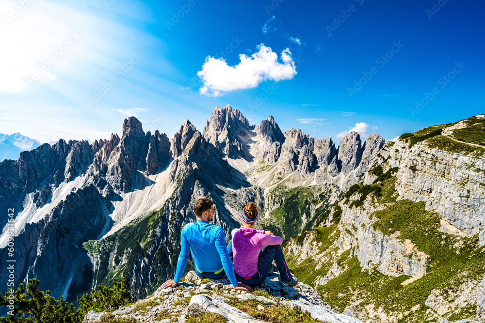 Fototapeta premium Sitting young couple enjoys epic view on Cadini di Misurina mountain range in the morning. Tre Cime, Dolomites, South Tirol, Italy, Europe.