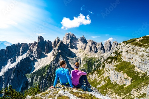 Sitting young couple enjoys epic view on Cadini di Misurina mountain range in the morning. Tre Cime, Dolomites, South Tirol, Italy, Europe.