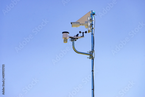 Security video camera and wind indicator against sky.