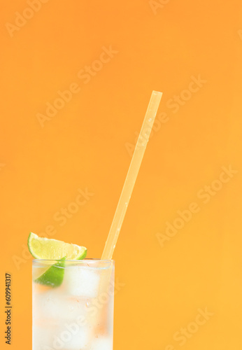 Lemonade with ice and lime on a bright orange background. Summer cocktail with cold drink. Copy space. Drink with a straw close-up