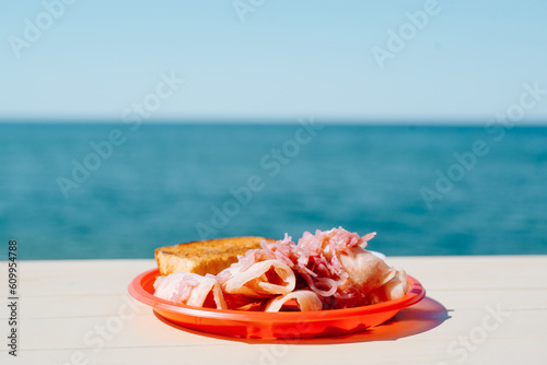 Close-up of dish of bonito stroganina in plate on table against backdrop of sea on sunny summer day. Sliced frozen fish with red onion and bread photo