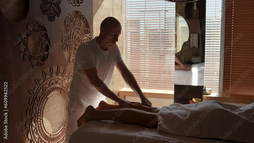 Remote view of masseur male giving essential oil massage of calf muscles to unrecognizable man lying on massage table in luxury spa salon by window. Young guy getting feet massage treatment. Remote view of masseur male giving essential oil massage of calf muscles to unrecognizable man lying on massage table in luxury spa salon by window. Young guy getting feet massage treatment.