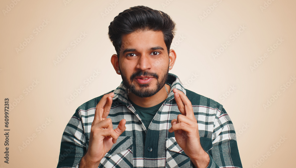 Indian young man crossed fingers looking at camera asking for good lucky news, wishing good exam results, dreaming about win victory in lottery jackpot. Hindu guy isolated on beige studio background Indian young man crossed fingers looking at camera asking for good lucky news, wishing good exam results, dreaming about win victory in lottery jackpot. Hindu guy isolated on beige studio background