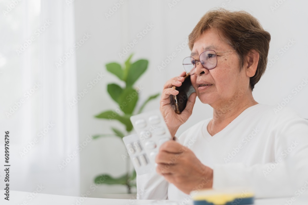 telemedicine and home health care concept, asian senior woman sitting at a table talking on a cell phone with doctor and holding a pill panel in her hand. telemedicine and home health care concept, asian senior woman sitting at a table talking on a cell phone with doctor and holding a pill panel in her hand.