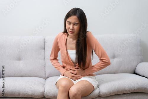 Sick Asian Woman Suffering From Acute Abdominal Pain in the abdomen due to menstruation period, PMS. Sitting On Couch, stomachache from food poisoning, abdominal pain, digestive problem, gastritis