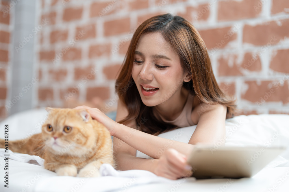 Beautiful Asian young woman using tablet and playing with her cat in the bedroom at home. Female relaxing with her cute pet. Beautiful Asian young woman using tablet and playing with her cat in the bedroom at home. Female relaxing with her cute pet.
