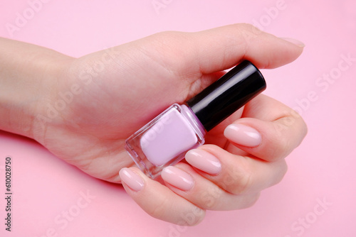 The hands of a young girl hold a bottle of pink gel nail polish on a pink background.The concept of hand and nail care, manicure, beauty salon, nail extension