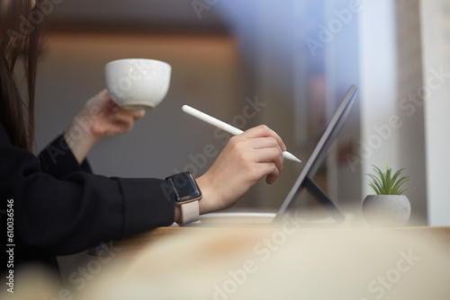 Professional graphic designer drawing a digital sketch on a tablet computer with a stylus pen. Freelance illustrator female drinking coffee and working in a cafe