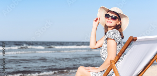 Happy brunette woman wearing sunglasses while relaxing on a wooden deck chair at the ocean beach.