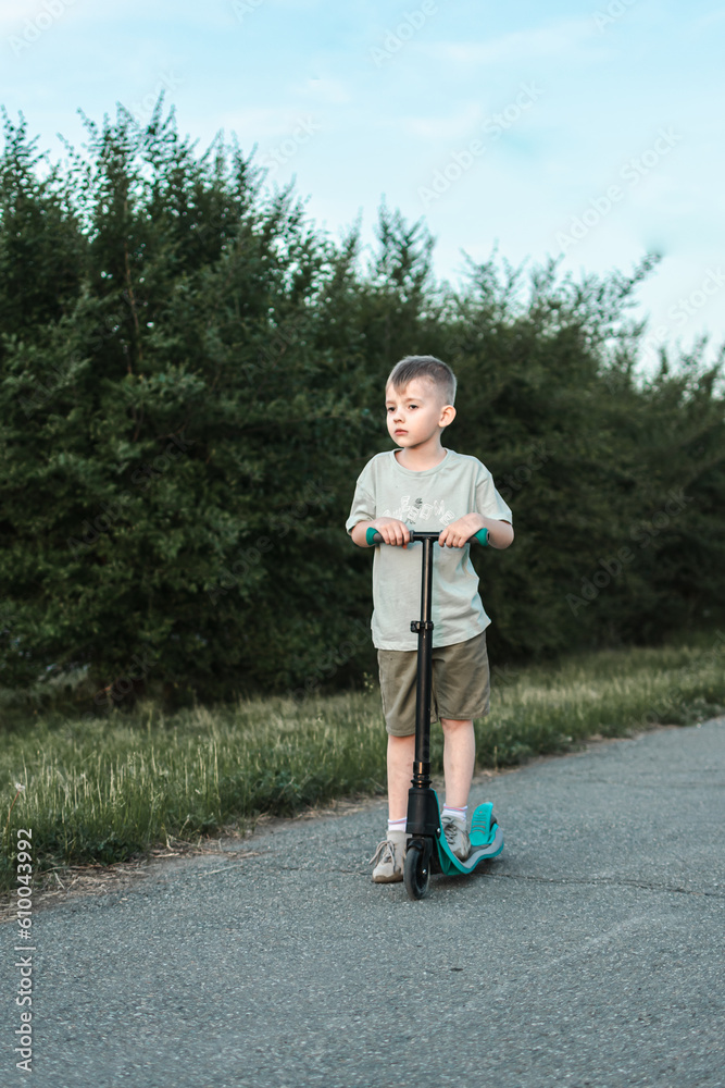 Fototapeta premium a boy in a light T-shirt rides a scooter along a path on a path covered with asphalt. children's portrait