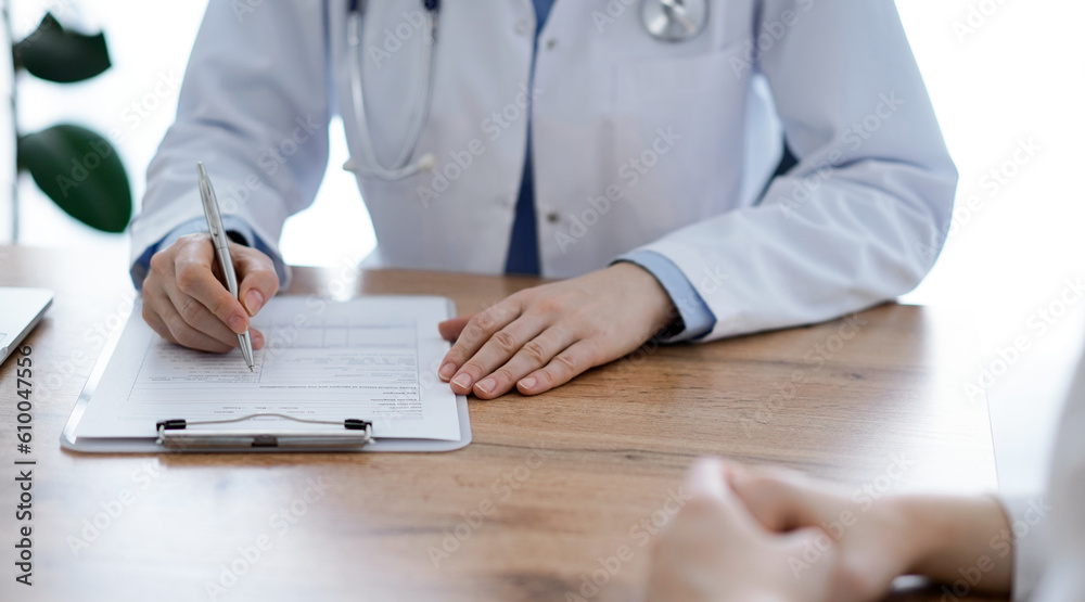 Doctor and patient sitting at the wooden desk in clinic. The focus is on female physician's hands filling up a records form. Medicine concept. Doctor and patient sitting at the wooden desk in clinic. The focus is on female physician's hands filling up a records form. Medicine concept.