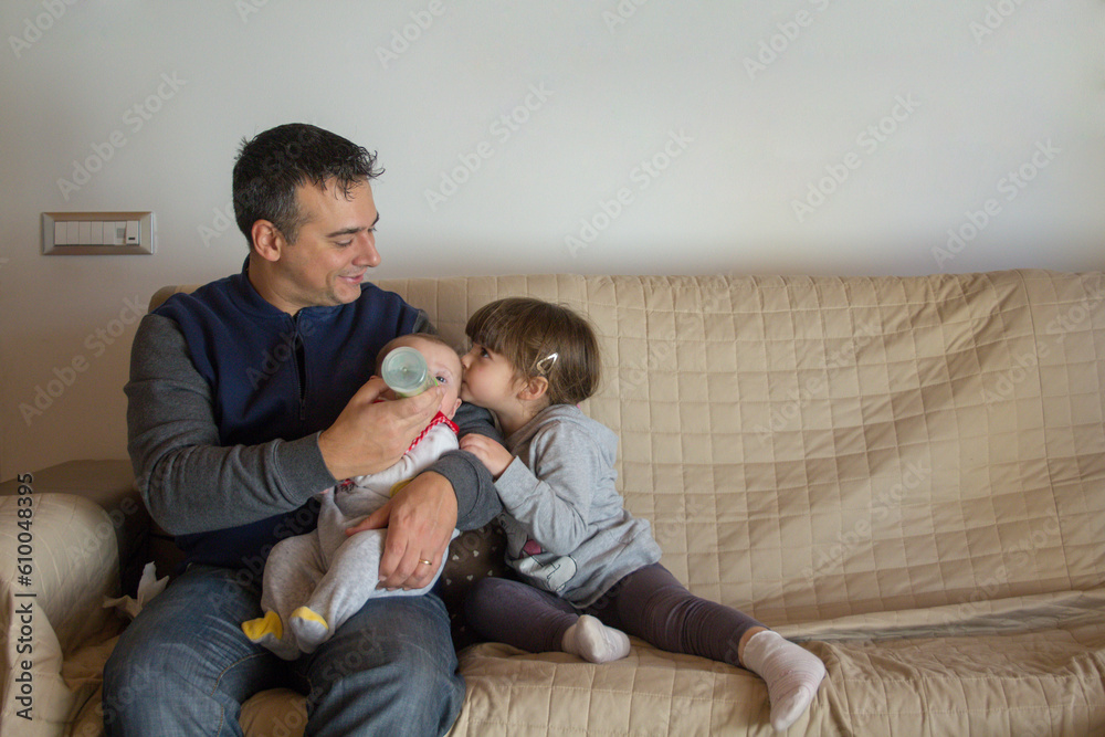 Image of a dad sitting on the sofa at home holding his newborn son in his arms while feeding him with the bineron and his older sister hugging and kissing him. Brotherly love and loving family
Image of a dad sitting on the sofa at home holding his newborn son in his arms while feeding him with the bineron and his older sister hugging and kissing him. Brotherly love and loving family