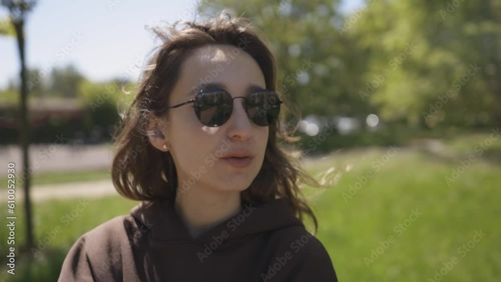 A Caucasian girl in dark sunglasses and a brown hoodie sits on a bench, an electronic cigarette in th park and holds a smartphone in her hand. The girl smokes e-cigarette and watching at smartphone. A Caucasian girl in dark sunglasses and a brown hoodie sits on a bench, an electronic cigarette in th park and holds a smartphone in her hand. The girl smokes e-cigarette and watching at smartphone.