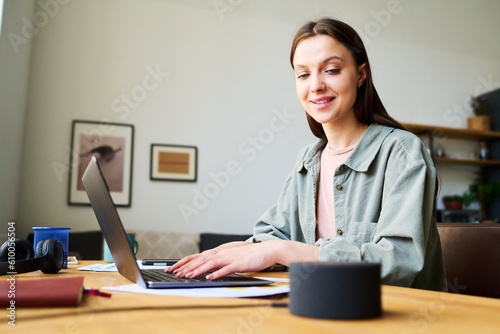Young woman talking to smart speaker while working online on laptop sitting at table in the room