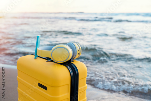 Yellow Suitcase with nd headphones on the  coast against the blue sea in amazing weather. Travel insurance and touristic concepts. Take music or book on a journey, #610062577