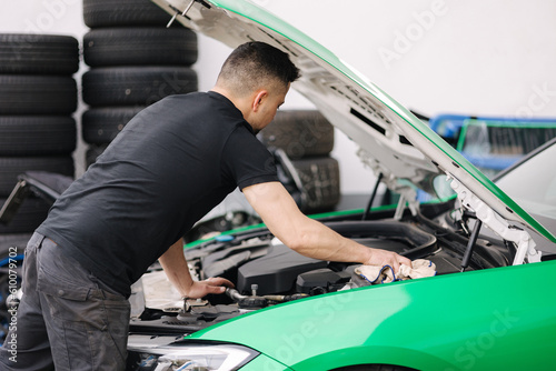 Portrait of a male mechanic working in his garage. Young bearded man checks the engine in the car, the hood is open