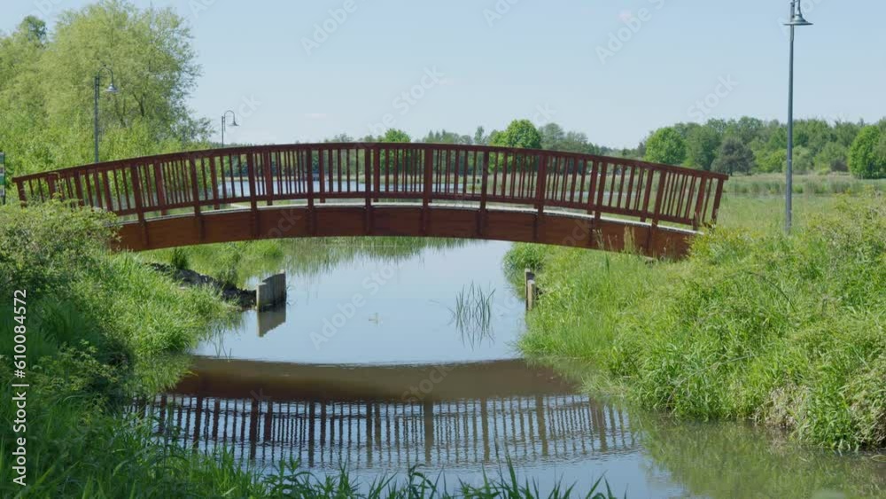 Wooden bridge over the river. Reflection of the bridge on the water. Landscape of a tourist place with a lake, a bridge, grass and lanterns. Trees in the background. Wooden bridge over the river. Reflection of the bridge on the water. Landscape of a tourist place with a lake, a bridge, grass and lanterns. Trees in the background.
