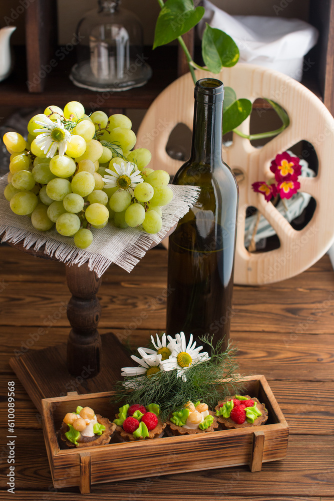 Obraz premium A bottle of wine, cakes and a branch of grapes on display. Wooden wheel in the background, Manor wine cellar