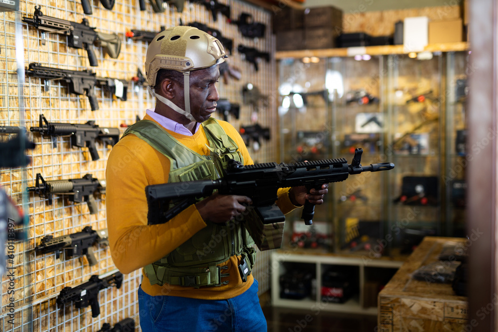 African-american man wearing military vest and holding machine gun in air weapon shop.