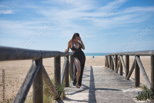 Attractive young woman, wearing black swimsuit and sarong, relaxed, solitary and calm, leaning on a railing of a wooden bridge, on the beach. Concept tranquility, peace, travel, vacation.