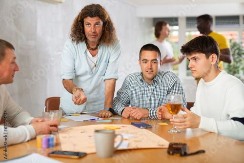 Joyous young and middle-aged men playing group table game with enthusiasm sitting at big desk © JackF