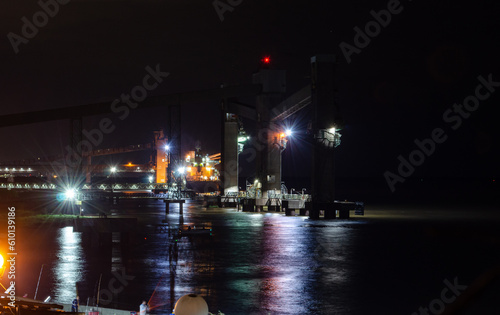 Night port scene with a cargo ship, little boat, fishermen on the pier, all on calm illuminated water.