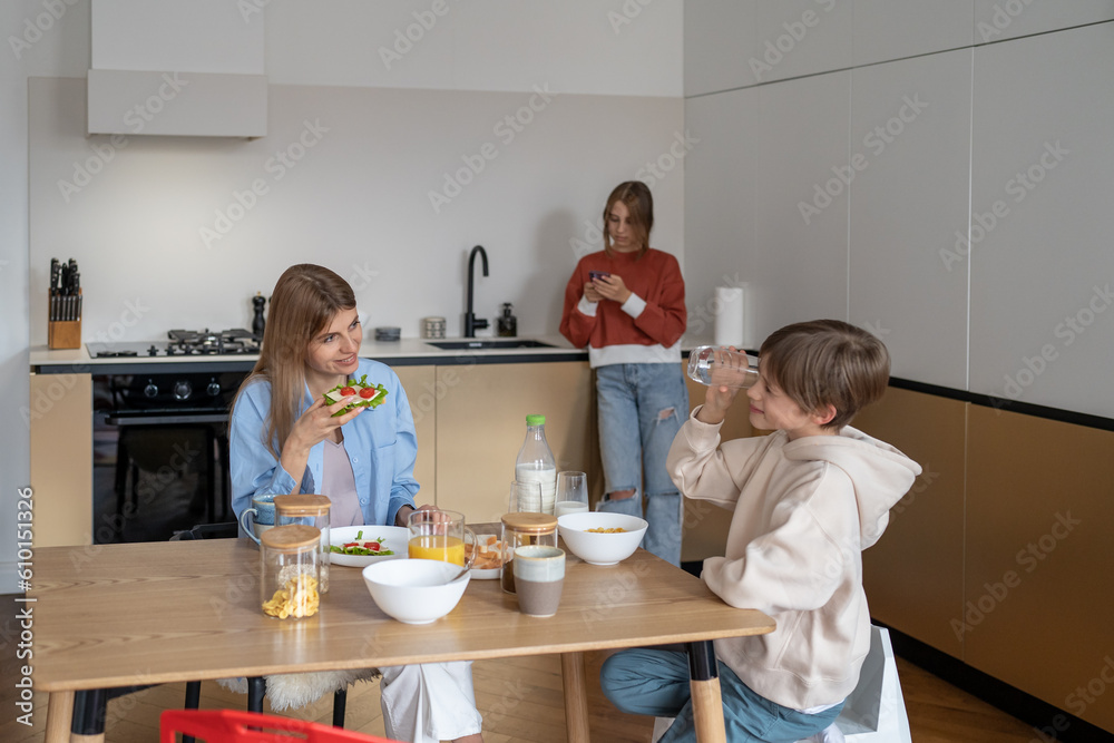 Family breakfast mom and kids. Young woman, teenager son eat at table in kitchen and talk. Eldest daughter stands aside and chats on phone in secret from mother. Relationship children and parents Family breakfast mom and kids. Young woman, teenager son eat at table in kitchen and talk. Eldest daughter stands aside and chats on phone in secret from mother. Relationship children and parents