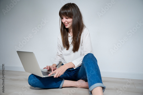 An attractive girl sitting on the floor works remotely on her laptop, responding to various messages and browsing the internet. Freelance work, remote learning, project work.